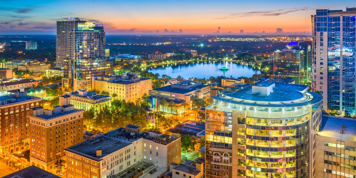 Nighttime aerial view of downtown Orlando, showing illuminated buildings and Lake Eola in the distance.